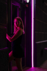 Club style photo of girl in a black dress.  Set is lit with violet light. Picture has dark night  tone. She stands at the entrance to the building