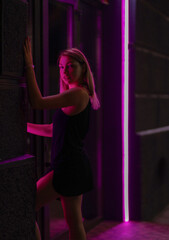 Fototapeta na wymiar Club style photo of girl in a black dress. Set is lit with violet light. Picture has dark night tone. She stands at the entrance to the building