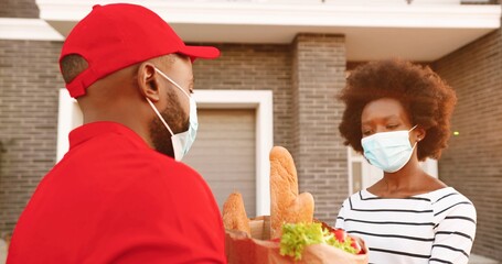 African American male courier in medical mask delivering bag with food to female client at house and handing outdoor. Delivery man bringing packet of products to woman customer. Corona lockdown.