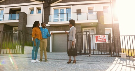 African American young woman real-eastate agent selling house to happy just-married couple. Outdoor. Wealthy man and woman buying dwelling in suburb. Businesswoman talking with new neighbors.