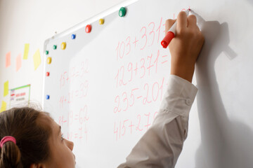 Small schoolgirl in a white blouse with pigtails writes with red marker on white board. Pupil...