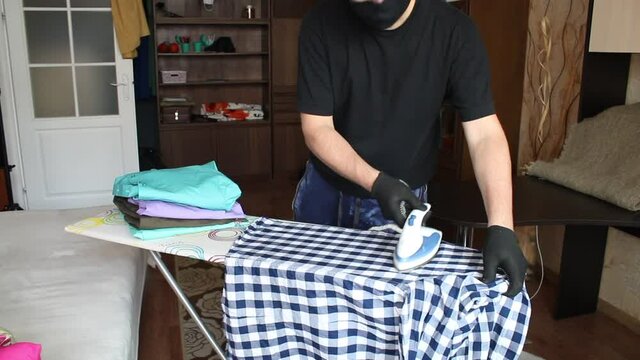 A man in a mask and gloves ironing a man's shirt. On the ironing board is a stack of men's shirts for ironing. Coronavirus protection