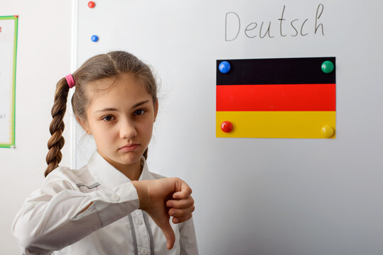Sad young student of secondary school in white uniform with pigtails is standing in front of board and showing thumb down. The flag of Germany on the blackboard. Concept of learning languages.