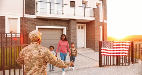 Happy African American woman wife with two small kids meeting man husband after army outdoor at big house in suburb. Male soldier coming back home to family and little children. USA flag.