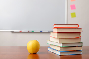 Stack of school books and a yellow apple on brown desk in a classroom, with white school marker board on a background. Free space.