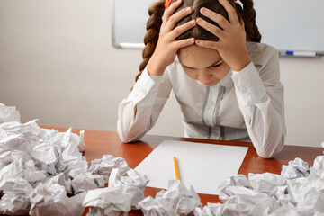 Irritated young schoolgirl with two pigtails sitting at the table with her hands around the head, thinking hard to find the ideas for writing essay, many crumpled papers around on the desk.