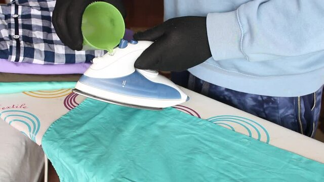 A man in gloves adds water to an iron. On the ironing board is a stack of men's shirts for ironing. Close-up shot.