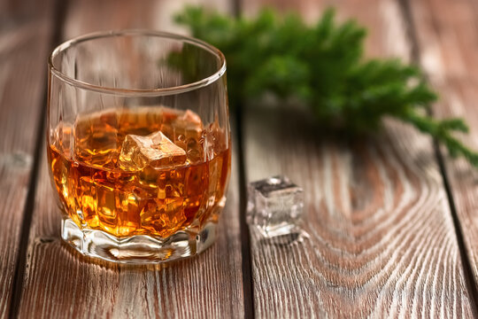 Whiskey with ice in a glass and a coniferous branch of a juniper on a dark old wooden table. Selective focus. Close-up