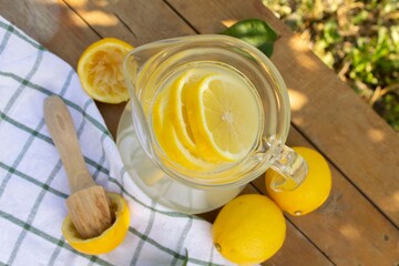 Top view of lemonade in a glass jug / pitcher with lemon slices. Lemons and wooden citrus pestle on wooden table with textiles outdoors
