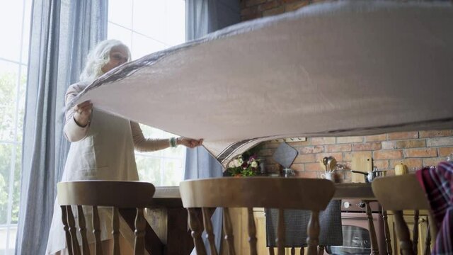 aged housewife in white apron covers large brown wooden table with tablecloth smiling and standing against orange brick wall and panoramic windows