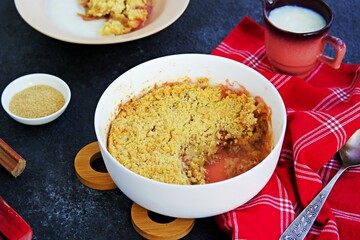 Dessert, crispy crumble with rhubarb in a white glass mold on a dark concrete background. Served with custard.
