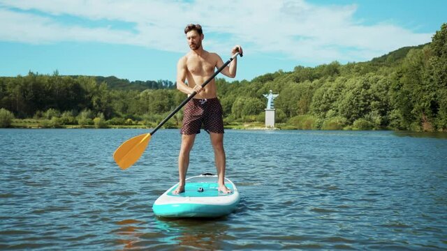 Handsome young man on sup paddle standing board at sunlight on lake summer water sport lifestyle travel boarding activity surfboard close up slow motion