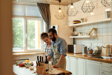 Fototapeta premium Young couple in love preparing the dough for making pizza with vegetables at home. Man and woman wearing apron, cooking together indoors. Hobby, lifestyle
