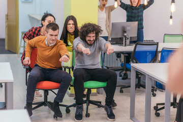 Team building and office fun.Four young cheerful businesspeople in smart casual wear having fun while racing on office chairs and smiling..