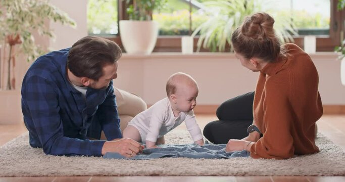 Smiling young couple lying together on rug on their living room floor at home with their adorable baby. Baby looks at mother and stands on all fours, mother and bearded father look at baby.