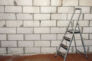 Construction stepladder on a wooden floor. Ladder near white building blocks wall, renovation. Copy...