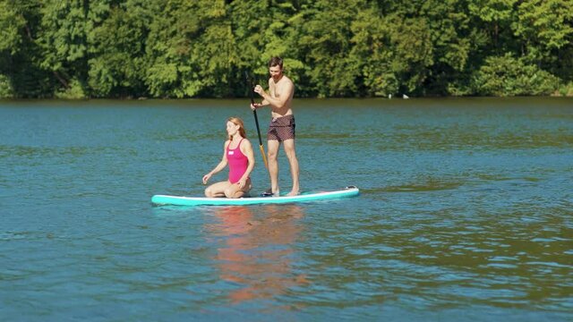 Together couple young man and woman in a bathing suit standing on sup board and swims surfing on the lake with a paddle rowing sport surfer friends close up slow motion