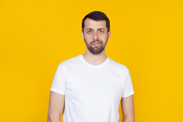 Young man with a beard in a white t-shirt happy face smiling looking at the camera. Positive man standing on isolated yellow background