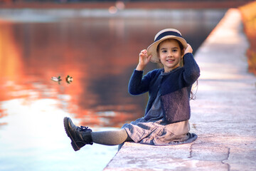 Fototapeta na wymiar A cheerful little girl sits on the shore of a pond,