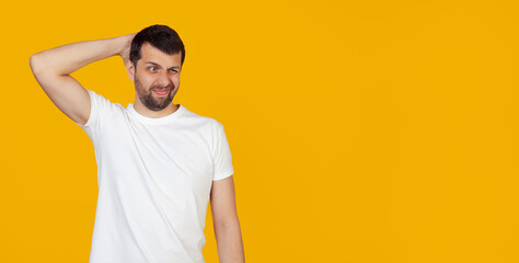 Young man with a beard in a white t-shirt worried about a problem, with a hand on his head, nervous and anxious about the crisis, stands on an isolated yellow background