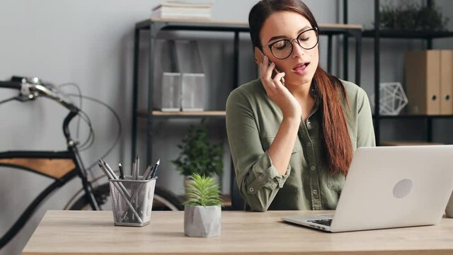 Portrait of young beautiful woman with glasses working on laptop. Female employee picks up phone and talking to client.