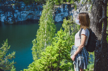 Young woman walking against the cliff with green trees and blue mountain lake wearing face protective mask for Covid 19 prevention. Traveler tourist with backpack. Corona virus and domestic tourism.