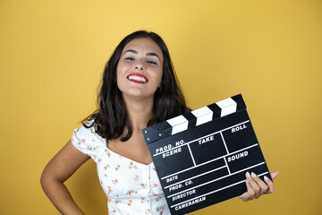 Beautiful woman over yellow background holding clapperboard very happy having fun.