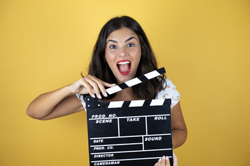 Beautiful woman over yellow background holding clapperboard very happy having fun.