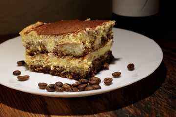 Slice of gourmet tiramisu surrounded by a sprinkling of coffee beans on a thin white plate atop a rustic wooden table with a bottle visible in the shadows behind. 