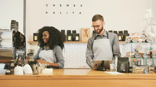 Barista woman and barista man working at bar table in cafe. Using coffee machine and digital device. African american woman. Caucasian man.