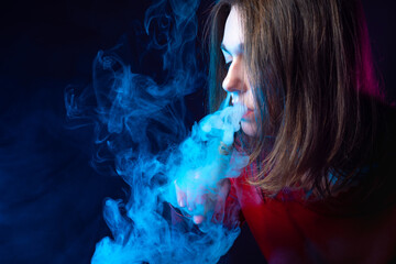 Portrait of a vaper girl in profile. A girl with an electronic cigarette in a cloud of smoke. A woman with an e-cigarette on a dark background with space for text. A long-haired girl smokes a VAPE.