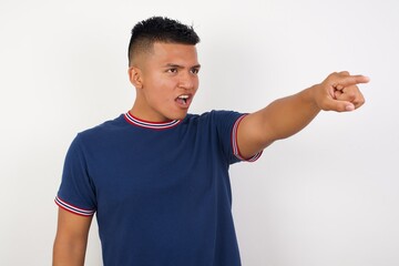 Young handsome hispanic man wearing casual t-shirt standing over white isolated background Pointing with finger surprised ahead, open mouth amazed expression, something on the front.