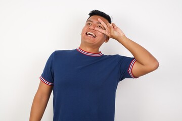 Young handsome hispanic man wearing casual t-shirt standing over white isolated background Doing peace symbol with fingers over face, smiling cheerful showing victory