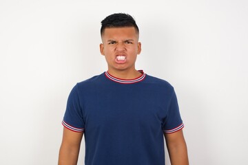 Fototapeta premium Young handsome hispanic man wearing casual t-shirt , keeps teeth clenched, frowns face in dissatisfaction, irritated because of much duties. Furious man looks out of window, sees miserable weather