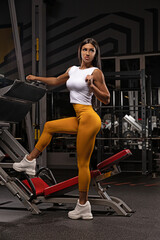 Fototapeta na wymiar A girl in the gym against the background of the simulator looks to the side and stands facing the camera on her topic, leggings and sneakers