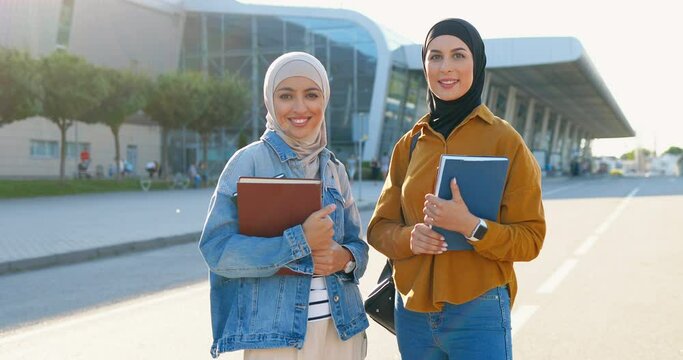 Portrait shot of young Arabic beautiful women in headscarves smiling and holding textbooks, books. Pretty Arabian females students in hijabs at street. Islamic happy girls. Education for muslims.