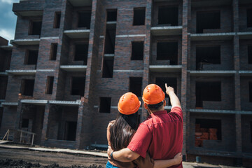 Back view a man and a woman in orange helmets stand with their arms around each other and look at a brick apartment building under construction. Investment in apartment, mortgage construction.