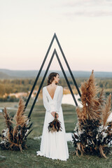 The bride with a bouquet of flowers stands in a triangular arch. Wedding ceremony in boho style.