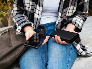 The girl uses a smartphone connected to the power Bank. Black smartphone and powerBank in women's hands close-up. Using gadgets in the modern world. The concept of portable chargers, modern tourism