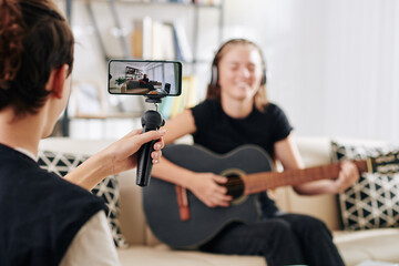 Teenage boy using smartphone on monopod when filming his sister or friend playing guitar and...