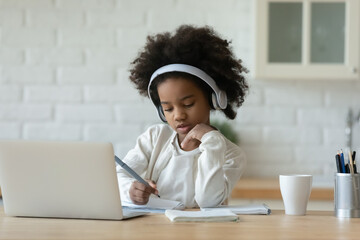 Focused little 7s mixed race child girl wearing wireless headphones, listening educational online...