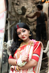 Obraz premium Portrait of stunning Indian woman draped in traditional saree adorned with gold jewellery, posing before the clay idol of the goddess Devi Durga