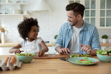 Happy multiracial family sitting at wooden countertop, enjoying cooking healthy food together in kitchen. Smiling little african ethnicity kid girl chopping vegetables with foster caucasian father.