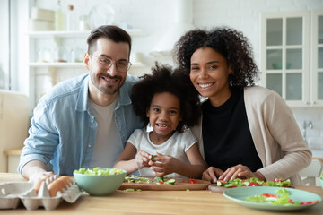 Portrait of happy mixed race couple with small girl cooking in kitchen. Smiling young handsome...