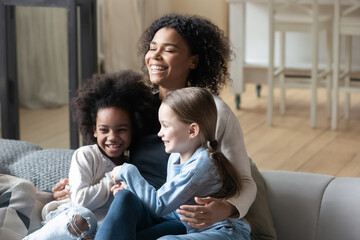 Happy young african american woman relaxing on sofa, cuddling playful diverse small mixed race...