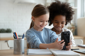 Happy little adorable multiracial children girls using smartphone application, sitting at table....
