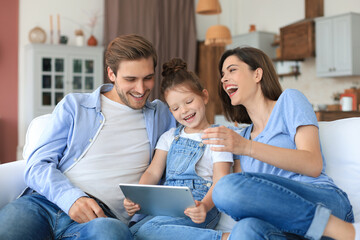 Positive friendly young parents with smiling little daughter sitting on sofa together answering...