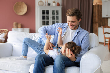 Smiling young father lying on couch at living room and play with happy little daughter.