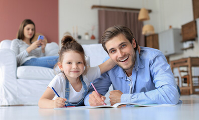 Obraz premium Happy father smilling daughter lying on warm floor enjoying creative activity, drawing pencils coloring pictures in albums, mother resting on couch, family spend free time together.