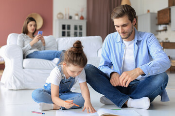 Obraz premium Happy father smilling daughter lying on warm floor enjoying creative activity, drawing pencils coloring pictures in albums, mother resting on couch, family spend free time together.